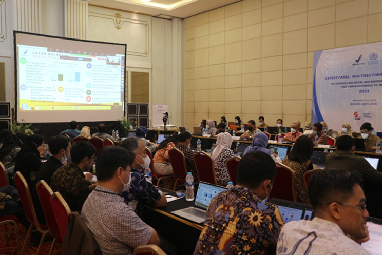 A conference room with participants seated at tables with laptops, facing a presentation screen displaying a slide with graphs and bullet points. Attendees appear engaged, some looking towards the screen and others at their computer screens.